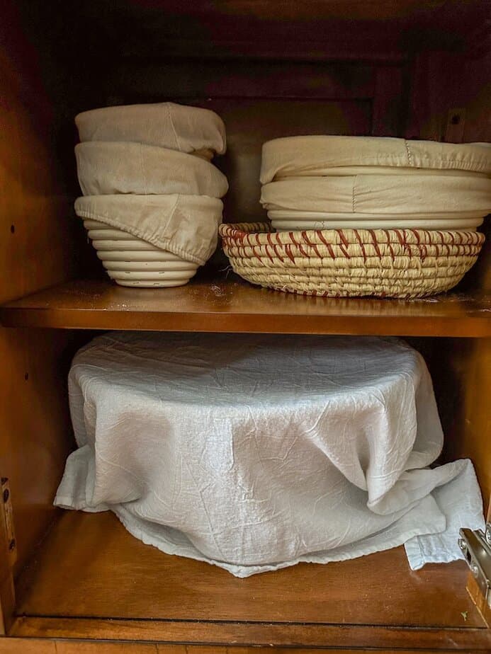 Wooden cabinet with banneton baskets on top shelf and a large bread bowl covered with a white flour towel on the bottom shelf.