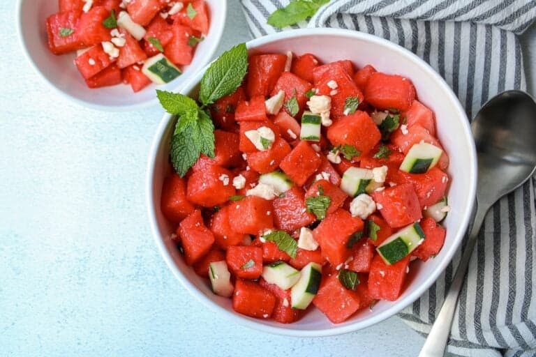 Cubed watermelon, cucumber salad with crumbled feta cheese and chopped mint in a white bowl.