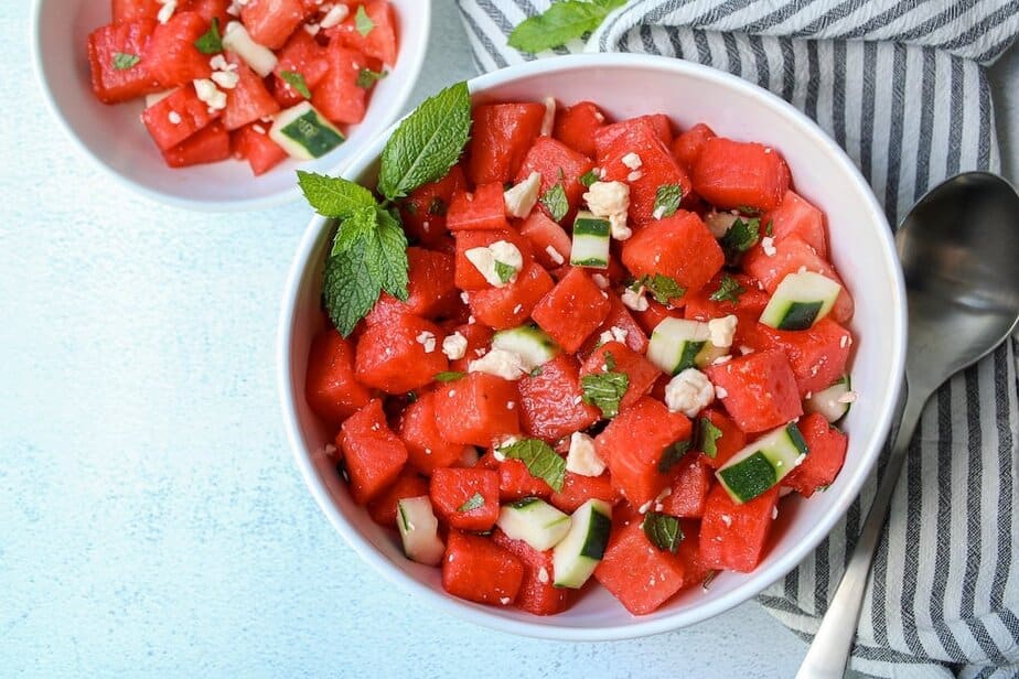 Cubed watermelon, cucumber salad with crumbled feta cheese and chopped mint in a white bowl.