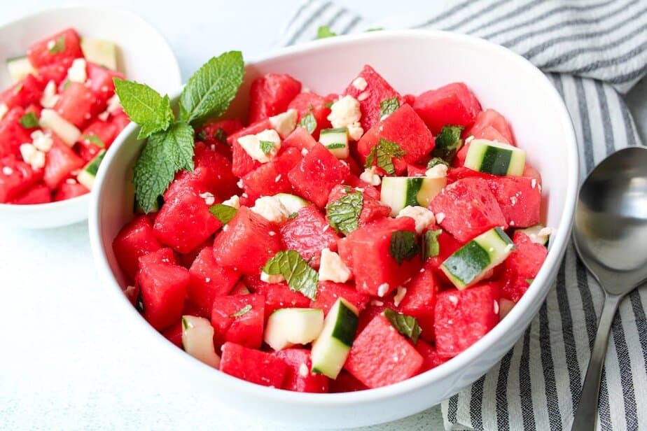 Cubed watermelon, cucumber salad with crumbled feta cheese and chopped mint in a white bowl.