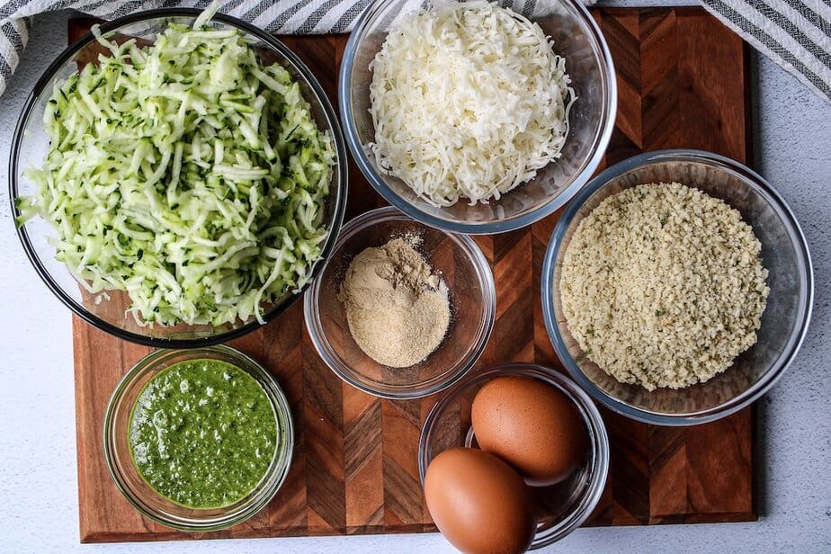 Ingredients in clear bowls: shredded zucchini, parmesan cheese, breadcrumbs, spices, pesto and two eggs.