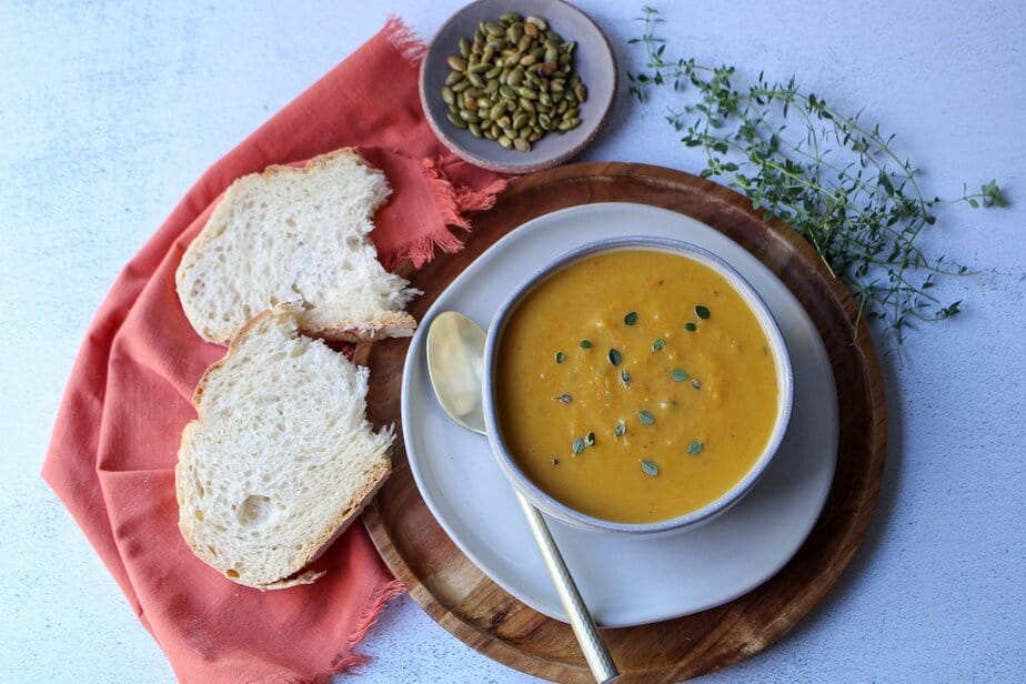 Creamy pumpkin soup in a bowl with bread, pumpkin seeds and herbs on the side. 
